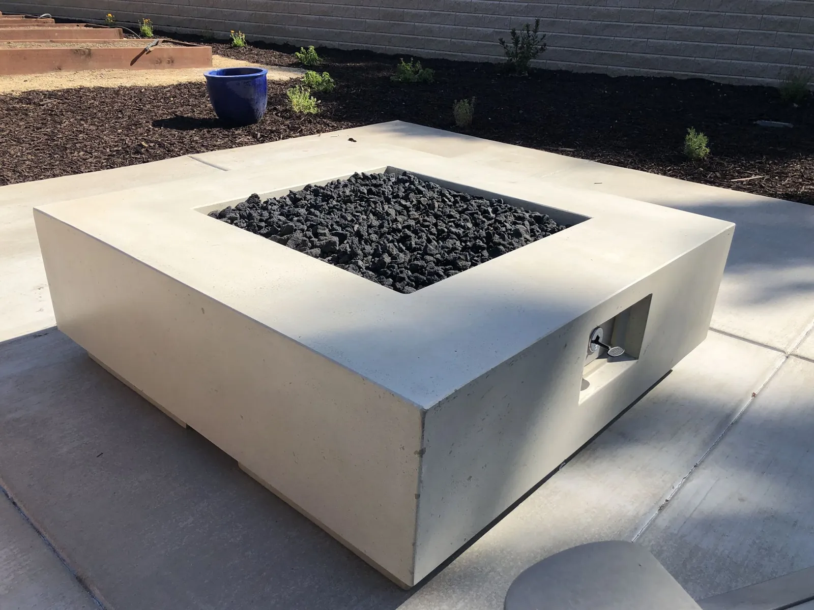 Square cast concrete fire table filled with black lava glass on a smooth concrete patio