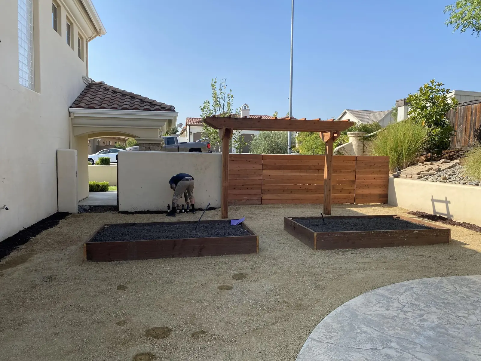 Raised garden beds and a pergola beside a stamped concrete patio and wood fence