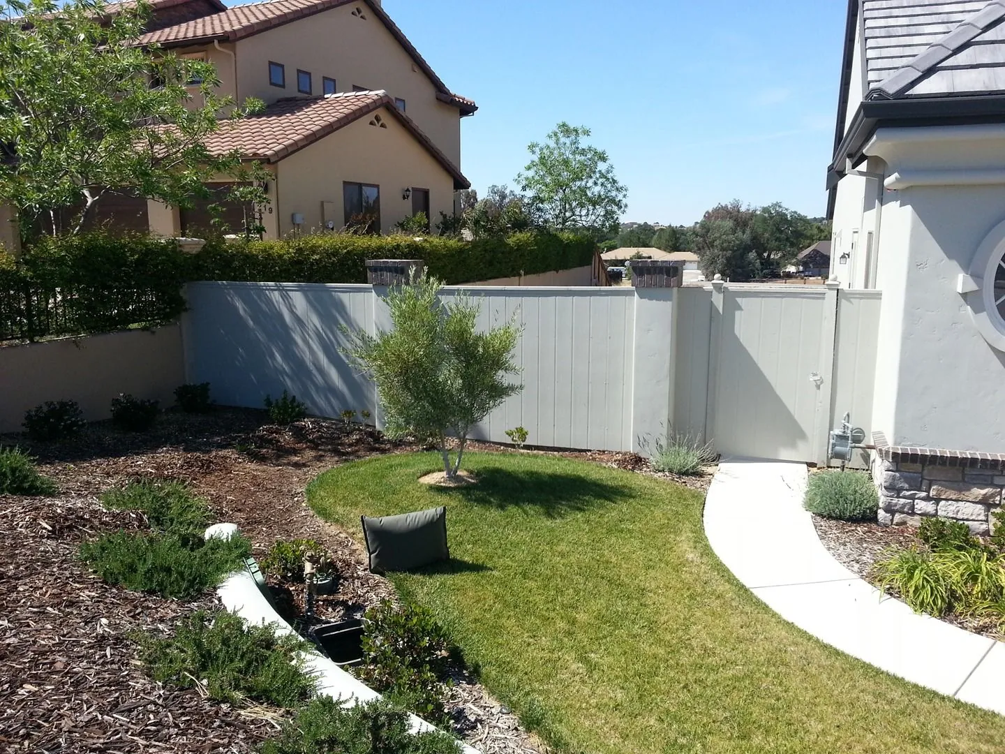 Curving concrete path along a side yard with a small lawn, mulched planting beds and a white vinyl fence