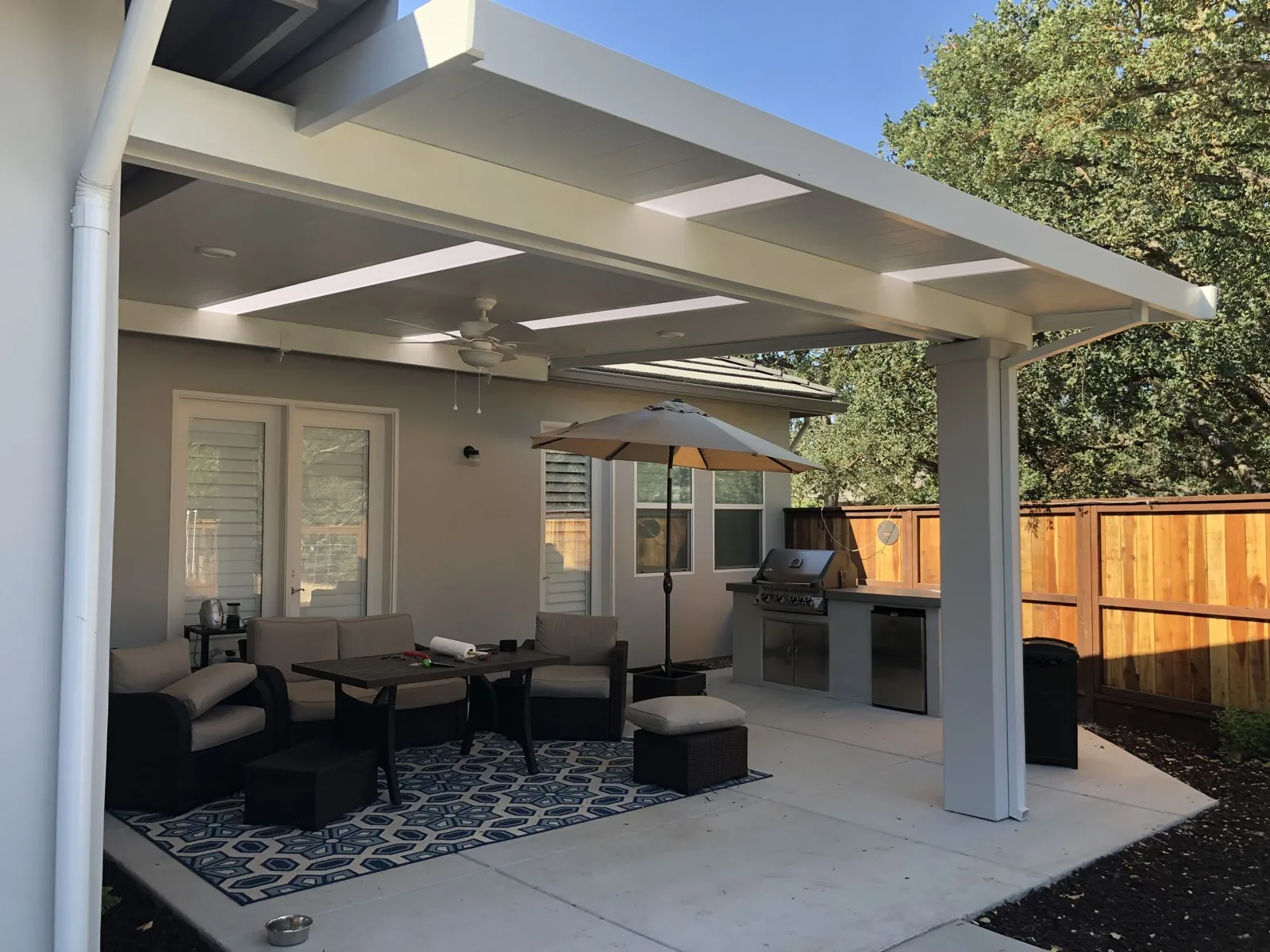 White aluminum patio cover over a concrete slab furnished with lounge seating and an outdoor kitchen beside a cedar fence